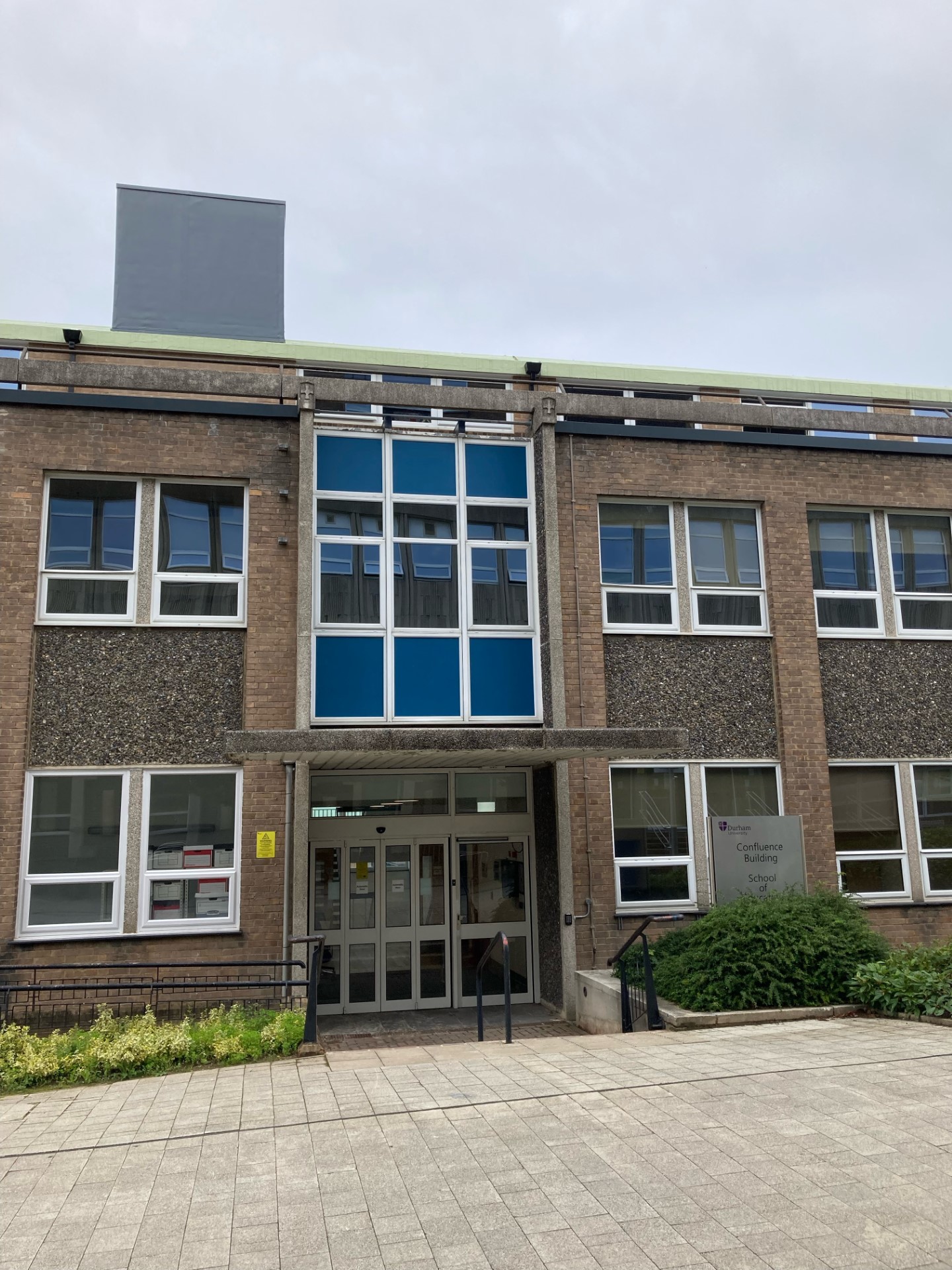 Photograph of brown-bricked 1960s building with light green roof, lots of windows and a pair of automatic doors in the centre.