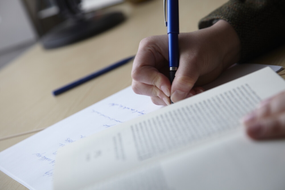 A student writing on a sheet of paper with a book open in the foreground