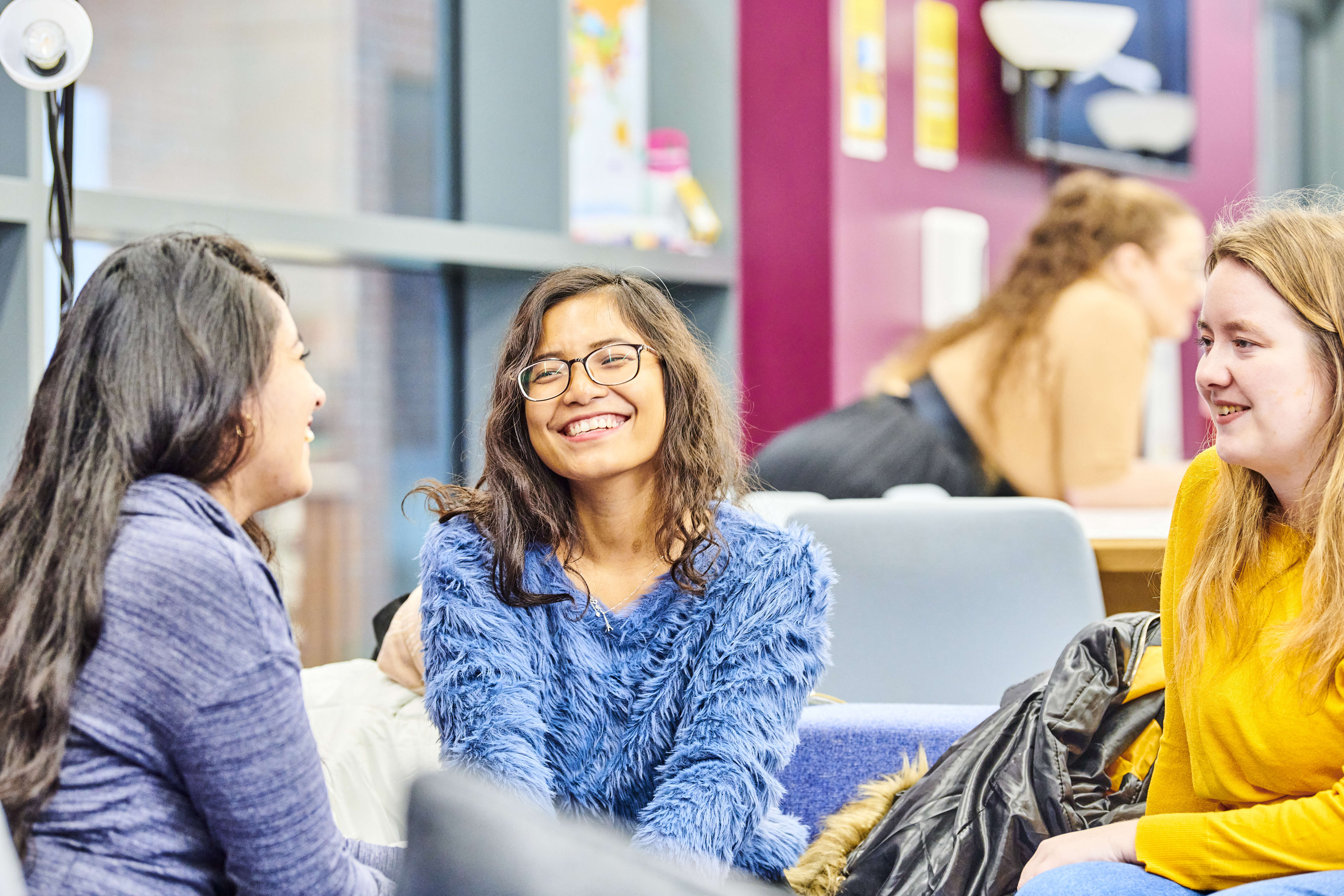 A group of female post-graduates sit together chatting and laughing. The space is brightly lit and colourful.