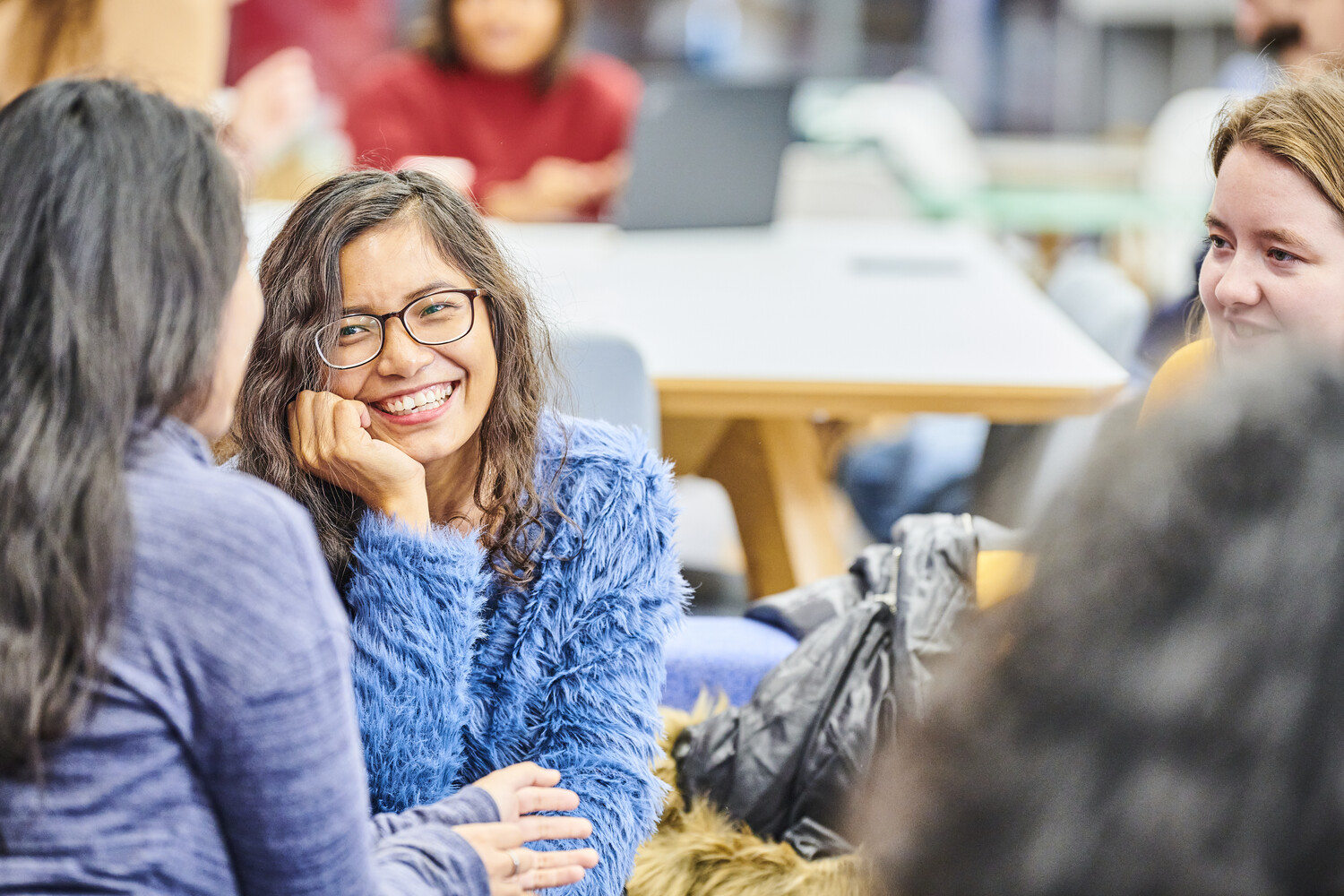 A group of female post-graduates sit together drinking coffee or tea, chatting and laughing. The space is brightly lit and colourful.