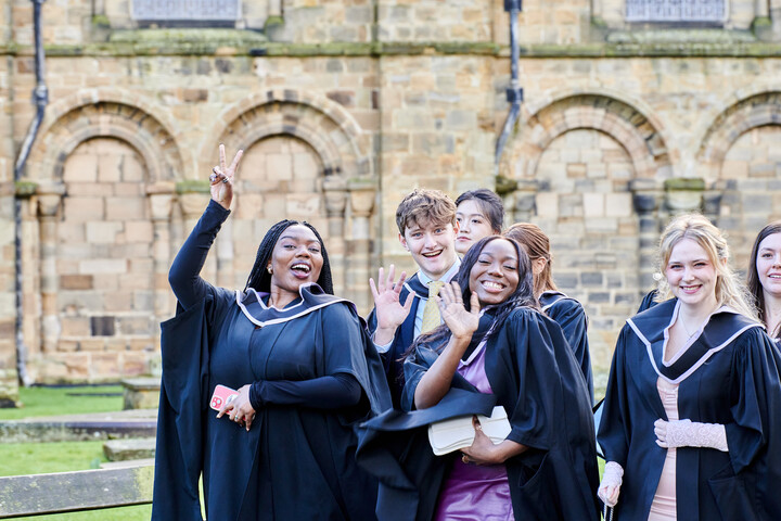 A group of graduates in their gowns, smiling and waving at the camera outside of Durham Cathedral