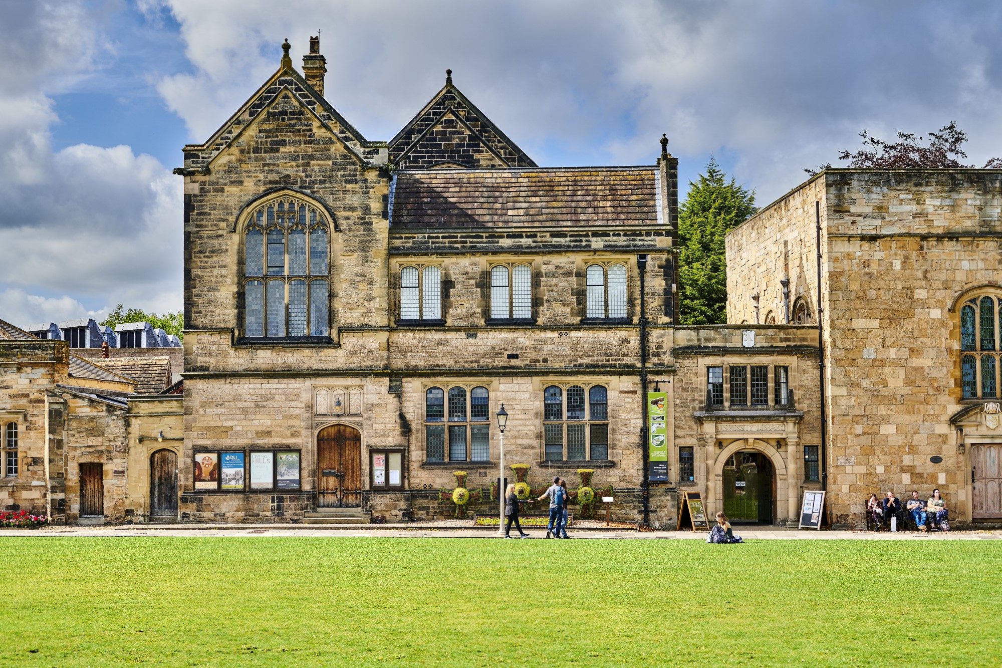 Series of historic brick buildings behind a large lawn, a group of people walk past while others sit on a bench in front of the building.