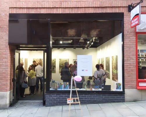 Looking into a large shop window from outside, inside the shop is an art exhibition on the walls, groups of people stand inside the shop chatting. Outside the shop is an A-board in side profile with a pink balloon attached to it.
