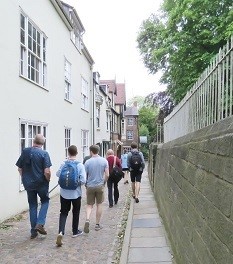 A group of people walk down a narrow, cobbled street, on the left are houses, to the right a wall with metal fence attached.