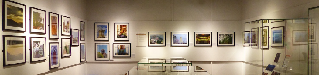 A rectangular gallery with white walls, black ceiling and red carpet. Around the walls are colourful photographs of landscapes in white mounts and black frames. Down the centre of the gallery are two display cases with many objects inside, another taller display case is visible on the right.