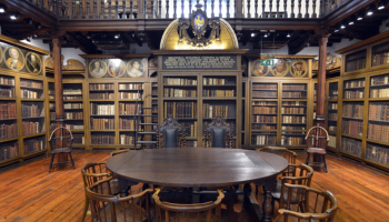 In the background, there are books on shelves with decorative detailing. In the forefront is a dark wood table with chairs around it.