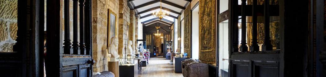 View along the length of the Tunstall Gallery inside Durham Castle. Along the length of the Gallery are tapestries, paintings, busts of famous figures, old wood and metal chests and museum cases.