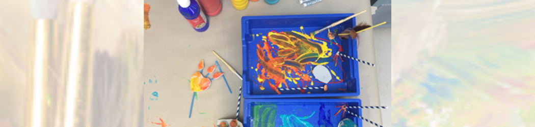 Downwards view of a table covered with bottles of paint, two blue trays filled with different colours of paint swirled together with paint brushes and straws lying in them and a completed artwork made of straws and card in the shape of a heart.