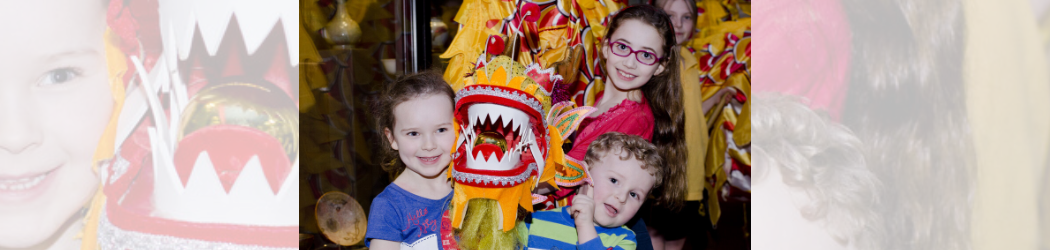 Four smiling children are holding up a brightly coloured Chinese parade dragon while standing next to a display case holding Chinese ceramics.