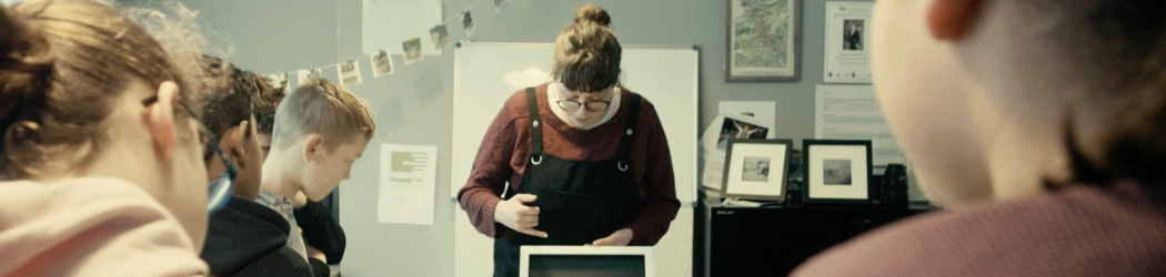 Photograph of an art display class – a member of the Learning and Engagement Team is holding up an artwork and pointing to it while talking about to a group of students who are gathered around it.