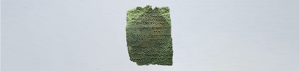 Photograph of a fragment of the Lanchester Diploma – a flat piece of corroded and damaged green metal covered in a Latin inscription that has been punched in and written in block capitals.