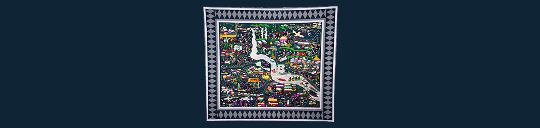 Photograph of a square fabric banner known as a story cloth. It is covered with scenes on a blue background showing the history of the Hmong people of South East Asia, who are depicted in traditional clothes of black with red belts. Traditional village life is depicted alongside images of forces migration from war and associated challenges.