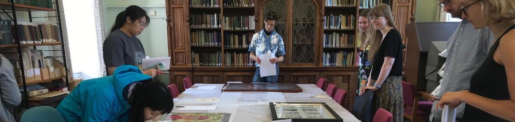 A group of students are standing around a table in the centre of the room, which is covered in bubble wrap. On top of this are framed images of artworks, which the group is looking at. One of the group has leaned over the table for a closer look.