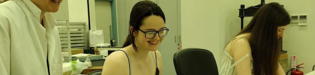 Two Work Experience participants sit at a table in a Conservation studio with historic documents and modern forms on the table. A member of the Conservation team stands next to them smiling.