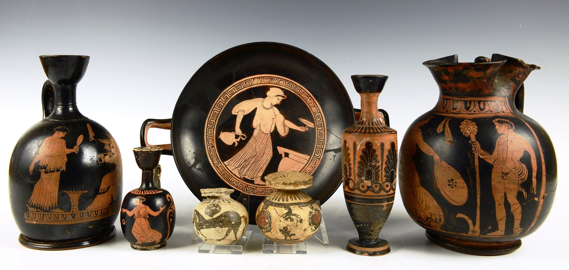 Seven pieces of Ancient Greek ceramics, on a graduated background. Ceramics include a wine cup, oil containers, and ointment pots.