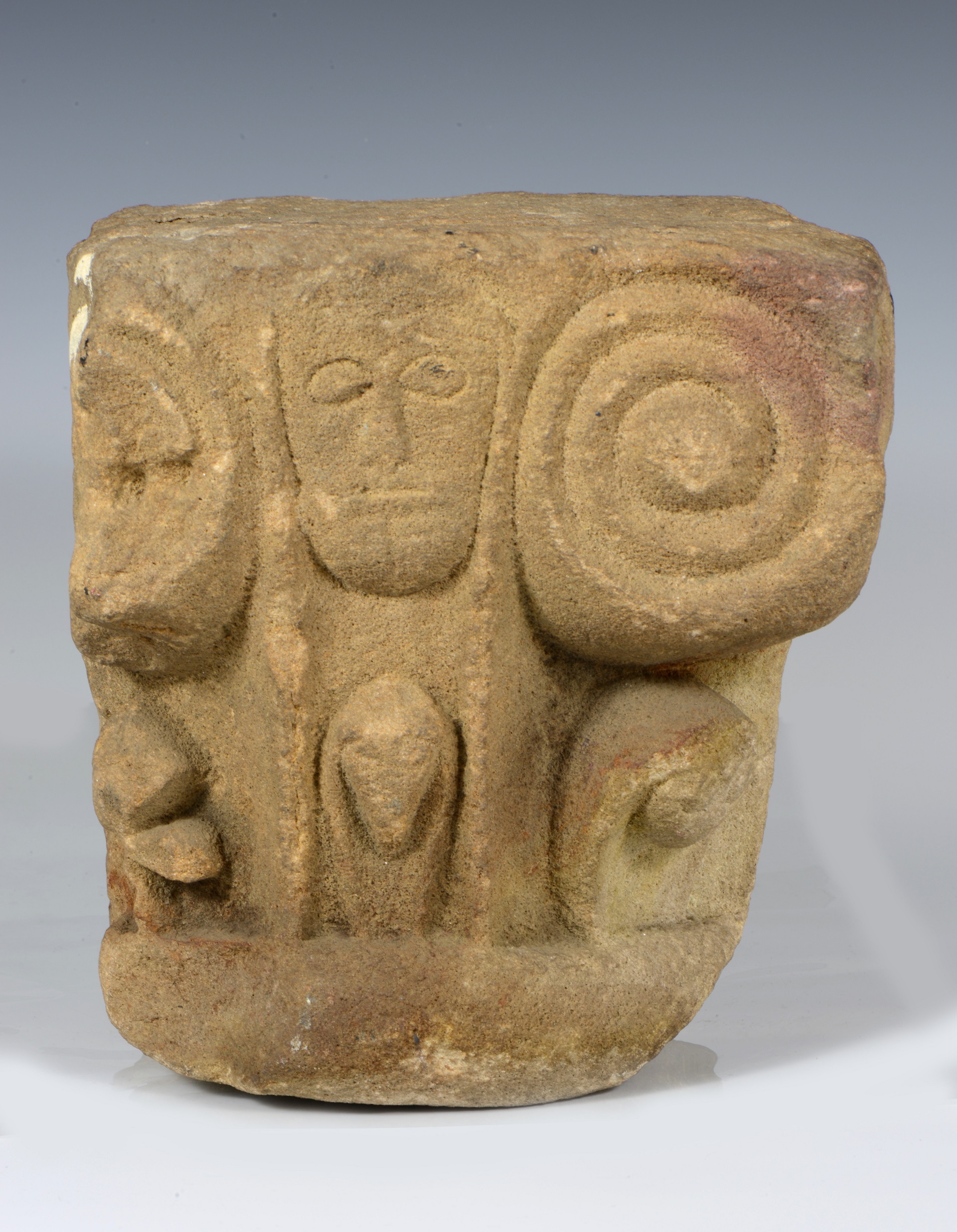 A piece of sandstone dating to the 12th century with carved decoration, including a face at the centre and scrolls on either side.