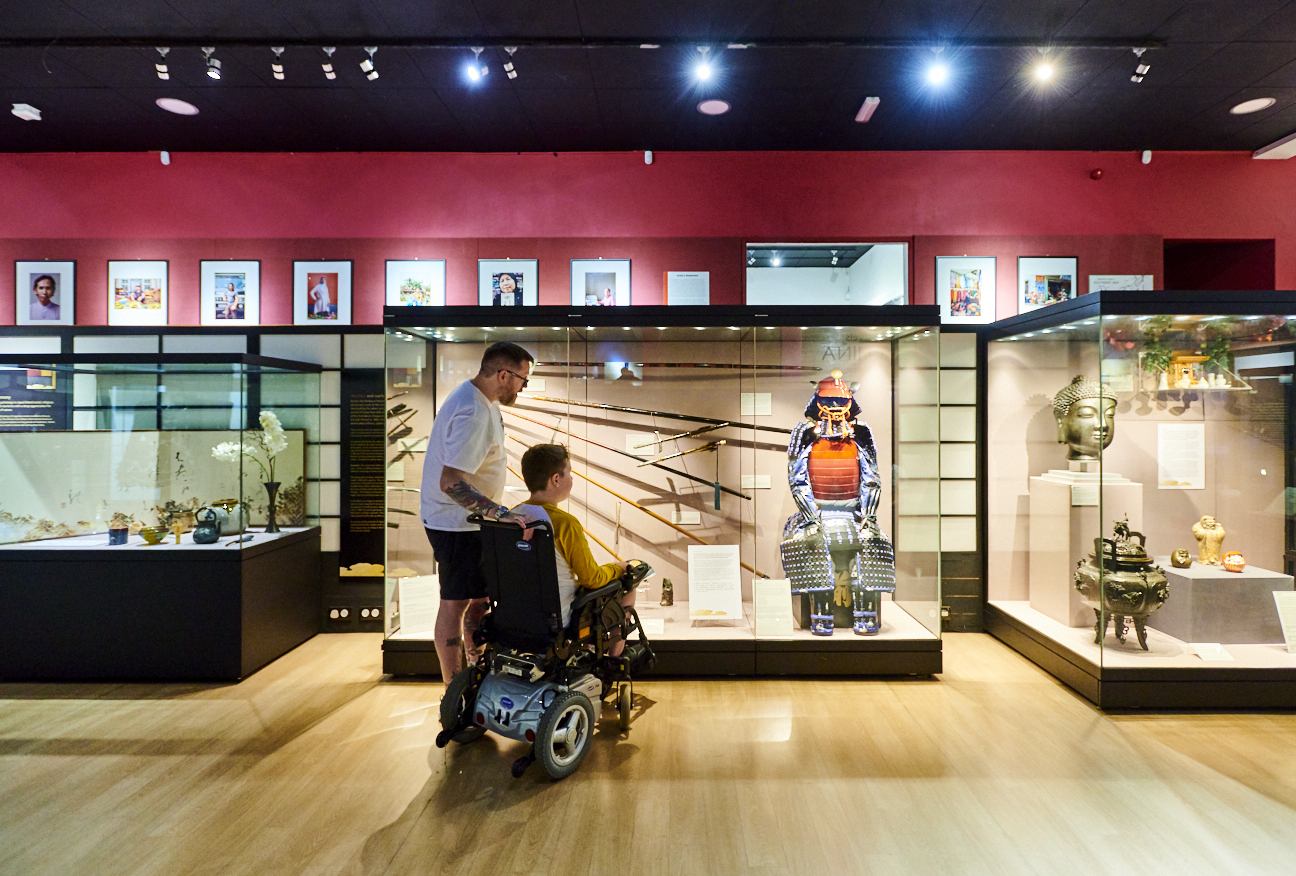 A man and a boy in a wheelchair look at displays of Japanese samurai weapons and armour, to the sides are displays of Buddhist art and objects relating to the tea ceremony.